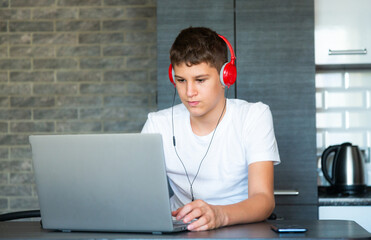 Cute young teenager in white shirt sitting behind desk in kitchen next to laptop and study. Serious boy in earphones makes homework, listening lesson. Home, distance education, self study by  kids.