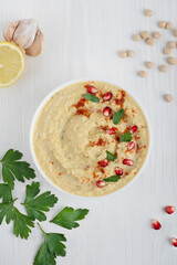 Wall Mural - Top view of organic vegan hummus thick spread made from mashed chickpeas, lemon and garlic used as a dip decorated with pomegranate seeds and parsley on white wooden background. Vertical orientation