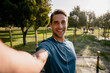 © Prins Productions - Smiling young male athlete takes photo of self before exercising outdoors in the sunny park listening to music in ear phones