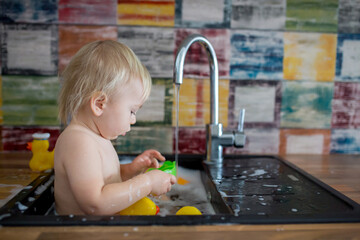 Canvas Print - Cute smiling baby taking bath in kitchen sink. Child playing with foam and soap bubbles in sunny kitchen with rubber ducks