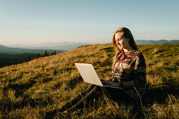 Hipster woman working with laptop sitting in mountains. Hiker tourist enjoying valley view sunset. Vacation holidays in autumn day. Distant remote work and travel, freelance as lifestyle concept.