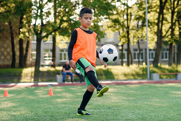 Motivated teen football player stuffs soccer ball on feet with boots. Practicing sport exercises at artificial stadium.