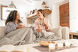 Two fine young women sitting on the sofa with a blanket on their legs laughing enjoying the house in winter having tea with pastries - Mixed race female couple home lifestyle