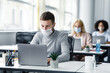 © Prostock-studio - Coworking office and safe work during coronavirus epidemic. Young man with face mask back at work in office after lockdown and looks at laptop