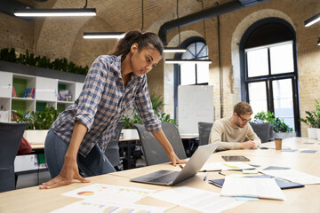 Wall Mural - Focused mixed race young business woman standing in the modern coworking space, looking at laptop screen and thinking about something while her male coworker working on the background