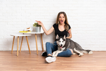 Young pretty woman with her husky dog sitting in the floor at indoors surprised and pointing finger to the side