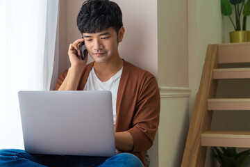 Prosperous young male freelancer making telephone call while working remotely on laptop computer beside window at home. Handsome smiling hipster guy enjoying mobile phone conversation.