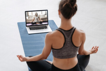 Close up of young sporty woman practicing yoga online with laptop at home. Yoga instructor conducting video training via video conference. Meditation, Sukhasana, relax. Healthy lifestyle concept