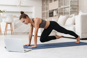 Young fit girl doing fitness exercise at home while watching online tutorial lesson on laptop. Beautiful athletic girl in leggings and top practicing yoga or workout in room. Sport, healthy lifestyle