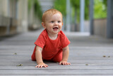 Fototapeta  - little happy baby in a red suit crawls on all fours on the floor in the park on a sunny summer day and smiles cheerfully, bokeh background, place for text