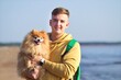 Portrait of happy young man, owner of beautiful Pomeranian spitz dog smiling at the sea, beach holding on hands, hugging cute little puppy