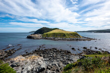 A Small Island Surrounded By The Blue Ocean. The Island Has A Grassy Knoll And A Small Hill Covered In Trees. The Sky Is Blue With Dramatic Clouds. There's A Steep Rocky Cliff With Beach Boulders.