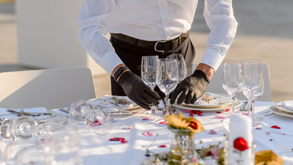 waiters hands in protective black gloves arrange a wedding party reception table decorated with flowers: plates, forks, knives and wine glasses.