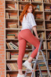 © oleg_ermak - Redhead female college student taking book from shelf in library. Stands on the stairs to get a book from the top. Side view, copy space.