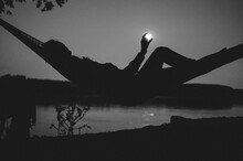 River Camping Under Full Moon; Silhouette Of A Girl In The Hammock Having Fun With The Moon