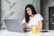 © F8  \ Suport Ukraine - Young woman typing on laptop and smiling while drinking juice in white kitchen