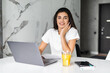 © F8  \ Suport Ukraine - Young woman typing on laptop and smiling while drinking juice in white kitchen