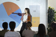 © Seventyfour - Side view portrait of young woman pointing at data chart with statistics while giving presentation during business conference, copy space