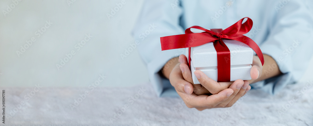 Obraz na płótnie Woman hand in a blue shirt holding a white gift box tied with a red ribbon present for the festival of giving special holidays like Christmas, Valentine's Day w salonie