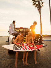 Boys (6-7, 10-11, 14-15) Waxing Surfboard At Sunset