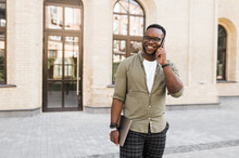 Smiling Successful African American Businessman Or Student In A Stylish Wear Talking On The Phone While Walking Down The Street And Holds A Laptop In Hand