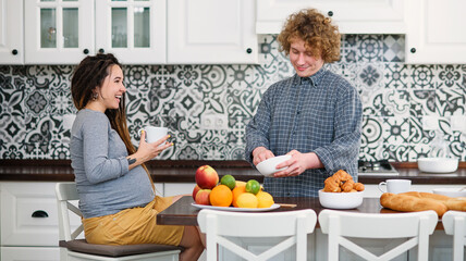 Joyful pregnant woman with dreadlocks drinks tea and watching her curly husband cooking breakfast for her in kitchen