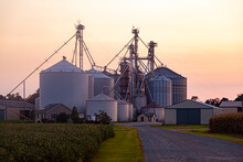  A Sunset View Of The Large Industrial Size Farm With Large Silos, Modern Metal Granaries And Granary Elevators. This Farm Specializes In Corn And Soy Bean Production