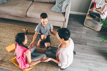 joyful family with positive brunette mother little son and teenager daughter plays funny game joining hands near soft sofa in light living room