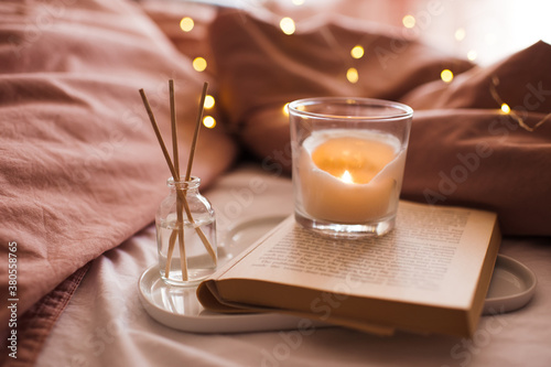 Fotografía Burning candle with aroma sticks in bottle on tray with open book in bed over glowing Christmas lights close up