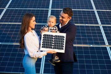 Wall Mural - Young happy family on the background of solar panels. A man, woman and baby boy are holding a solar panel in their hands and smiling. Solar energy concept image