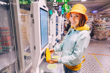 Happy young customer woman buys and pays for a bottle of orange juice at the checkout of a self-service vending machine in a modern supermarket, to avoid waiting in line