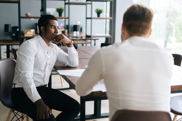 Thoughtful serious professional African American man interviewing caucasian European young candidate for job at successful company. Two business people talking strategy sitting at office desk.