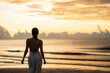 © marchsirawit - Woman in top bikini and white long pant on the beach with a beautiful sunrise or sunset in background.