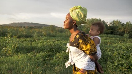 a tracking shot from the side of an african mother with her baby on her back walking by a lake in th