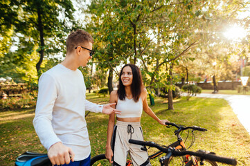 Positive young sports couple standing in the park with bicycles in the autumn season and talking with a smile on their faces. Happy man and woman resting after cycling in the park.