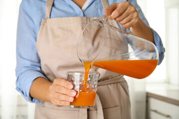 Wall Mural - Woman pouring freshly made carrot juice into glass in kitchen, closeup