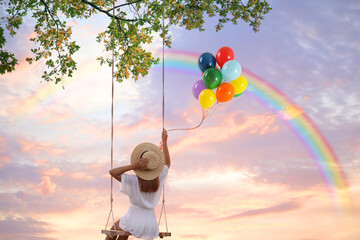 Dream world. Young woman with bright balloons swinging, rainbow in sunset sky on background