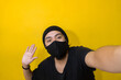 © exequiel - a young man waving at the camera, taking a photo or a selfie wearing a mask to protect himself from the virus, with yellow background