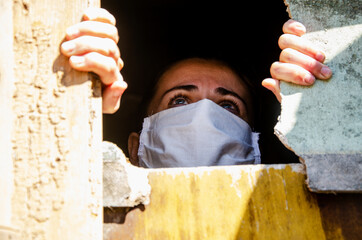 A girl in a medical mask on her face looks out the window close-up. Masked girl portrait photo. Mask mode during quarantine introduced due to coronavirus. People concept within coronavirus