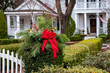 © Jen Wolf - Close-up of natural Christmas wreath of evergreens, berries, and red ribbon on an ivy-covered gate post at the entrance to the front garden of a beautiful white house.