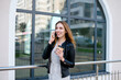 © Sergey - Beautiful woman is speaking on the phone and smiling, with a cup of coffee in the hand, against the glass facade in the city