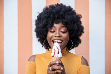 Close up shot of fashionable African American woman with curly hairstyle, eats delicious ice cream in waffle cone of strawberry flavor, wearing yellow turtleneck, posing on striped background.