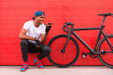 Full Body Of Cheerful Young African American Guy In Trendy Outfit And Cap With Earbuds Sitting Near Bike And Sending Audio Message On Mobile Phone While Resting Against Red Wall On Street