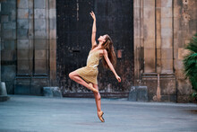 Full Body Of Graceful Young Female Dancer In Pointe Shoes Performing Elegant Dance With Arm Raised On Narrow Paved Passage Against Aged Stone Building