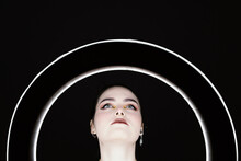 Female With Professional Makeup And In Stylish Wear Standing In Studio Behind Ring Circle Lamp On Black Background And Looking Away