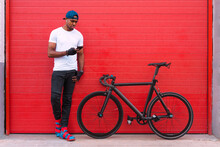 Full Body Of Young African American Man In Trendy Outfit And Cap With Earbuds Standing Near Bike And Sending Audio Message On Mobile Phone While Resting Against Red Wall On Street
