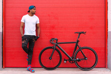 Full Body Of Confident Young African American Male In Street Style Clothing With Bicycle Leaning Back On Red Shuttered Door Of Urban Building