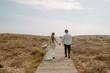 © Jake Jakab/ADDICTIVE STOCK - Full body back view of unrecognizable young newlywed couple in stylish clothes with bouquet holding hands and walking on wooden pathway towards sea
