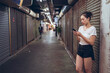 © Juan Alberto Ruiz/ADDICTIVE STOCK - Full body young Asian female in casual clothes with mobile phone in hand walking on narrow pedestrian underground pathway with closed shops in city