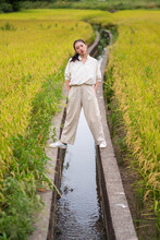 Calm Ethnic Female Standing Above Irrigation Canal Between Green Rice Fields And Looking At Camera